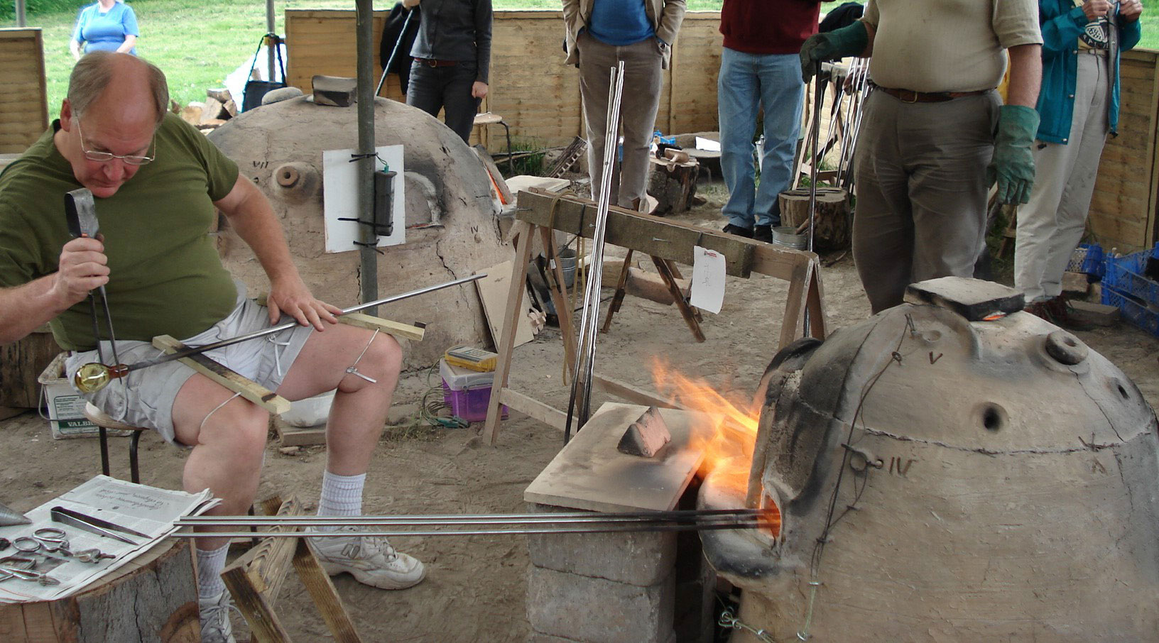 An individual in shorts and t-shirt using tools to shape glass on a blowpipe at a wood-fired furnace with onlookers in the background