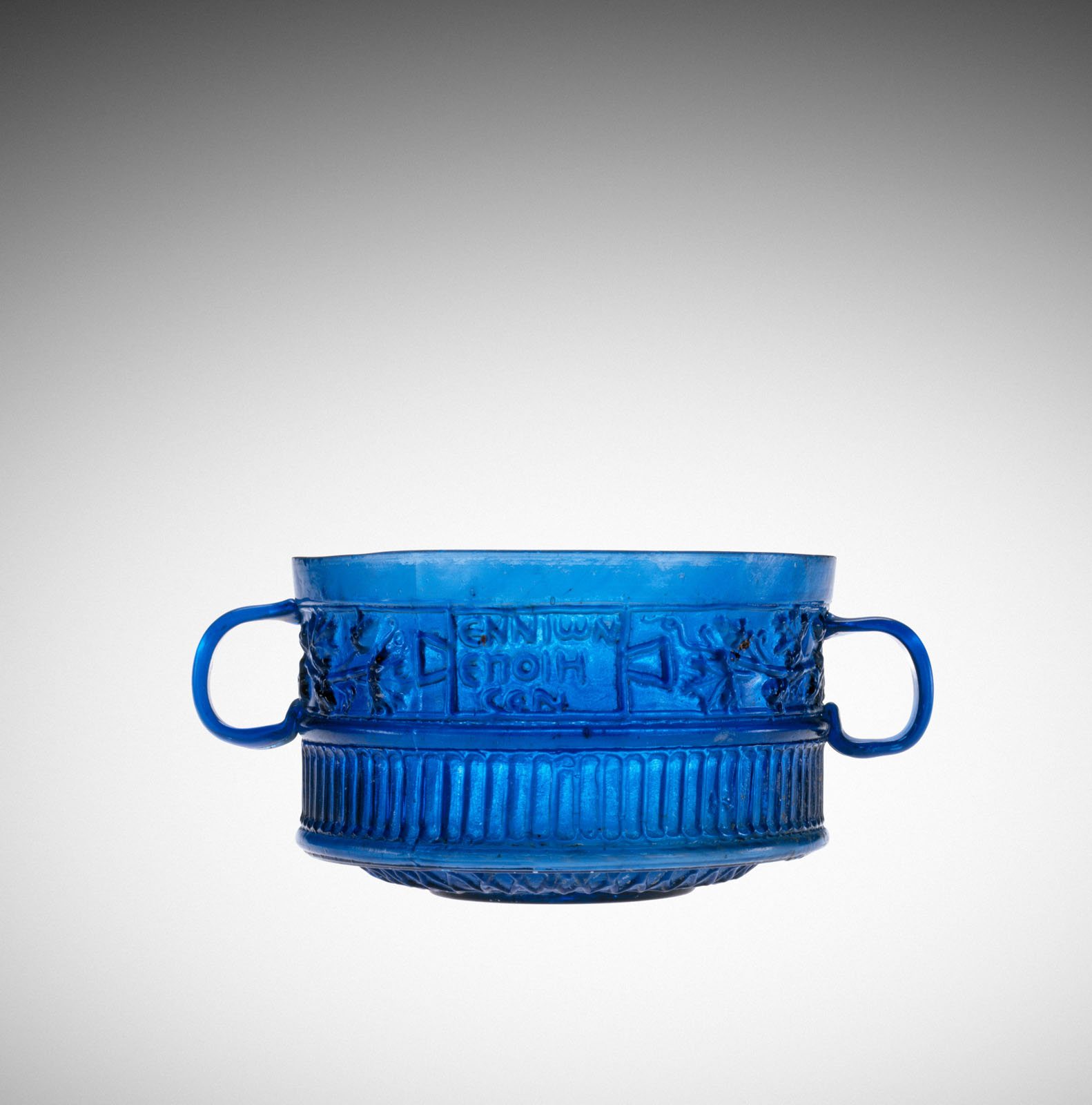 Vibrant blue glass cup with two handles, embossed with decorative details and writing in a band around its exterior