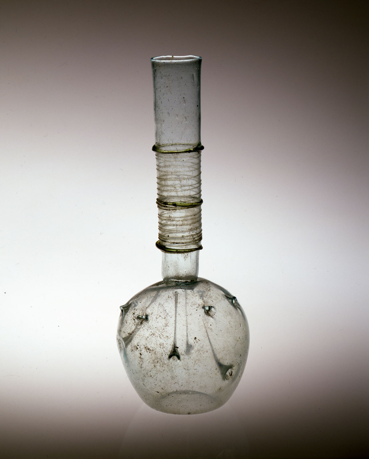 Transparent glass vessel with a long, narrow neck featuring spiral decorations and flattened, round base, showing signs of age