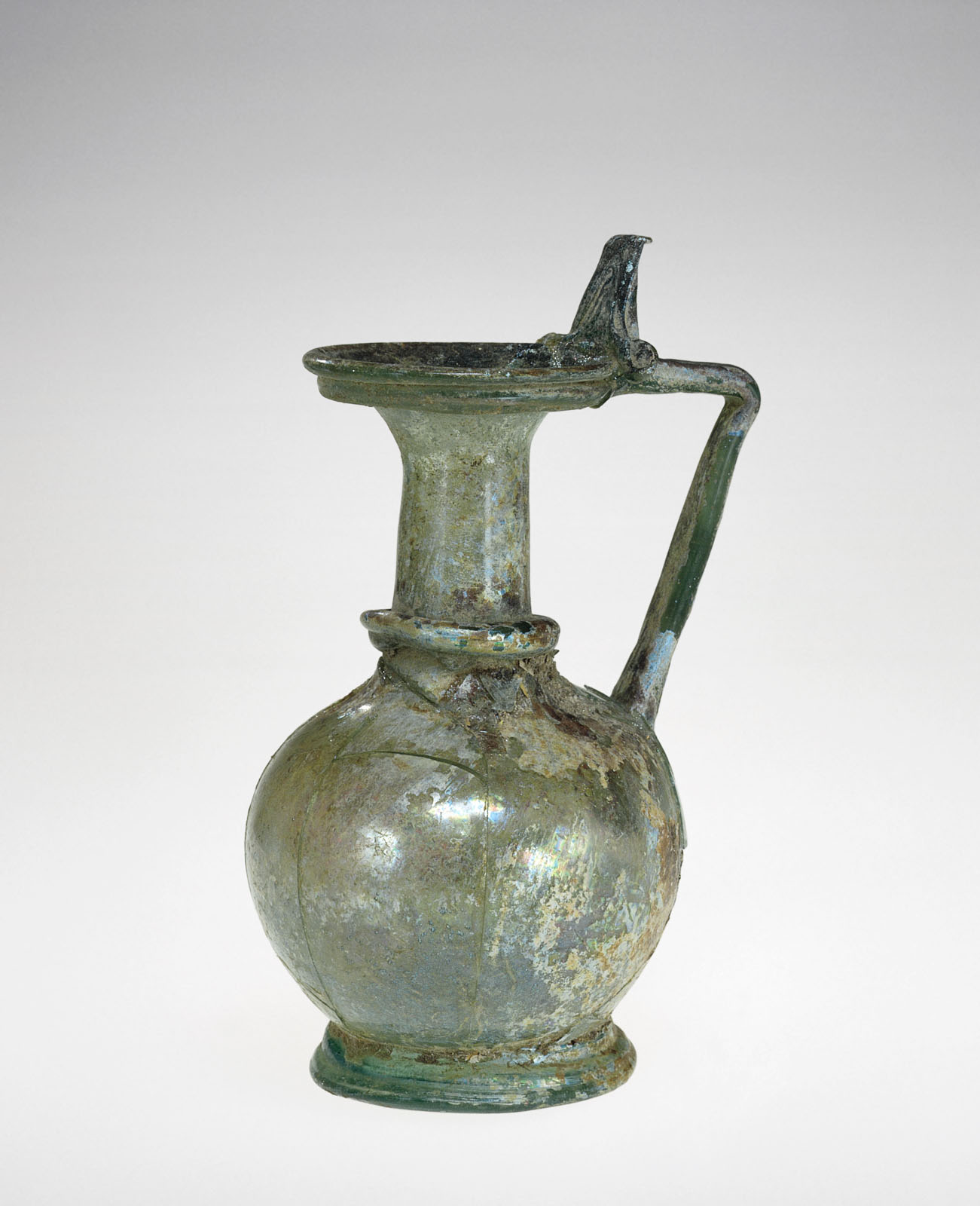 Mottled blue-green glass pitcher with a wide rim, narrow short neck, flat angled handle, and a round body, the surface showing iridescence from weathering