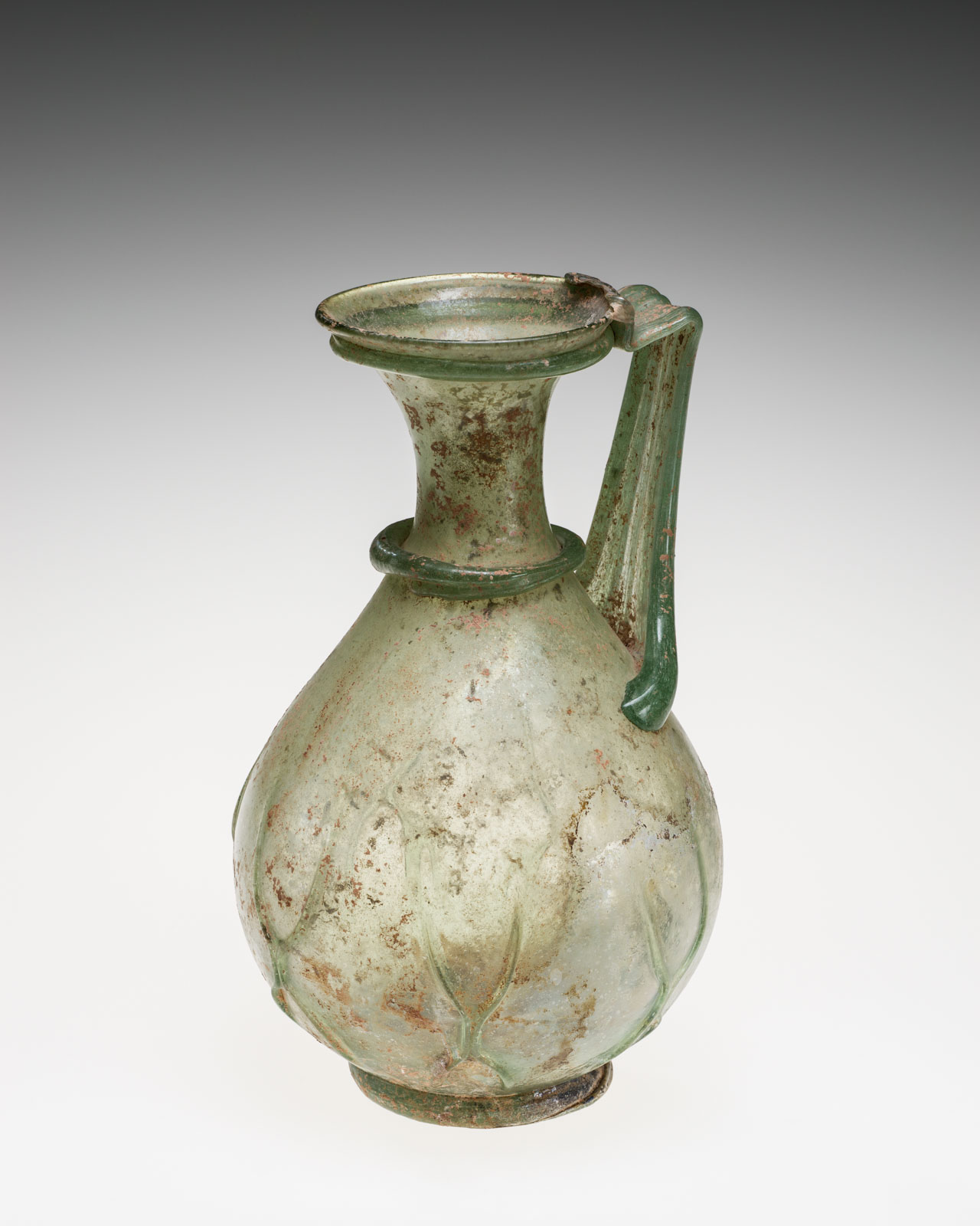 Pale green glass jug with rim, flat handle, and patterned bulbous body, showing a weathered texture