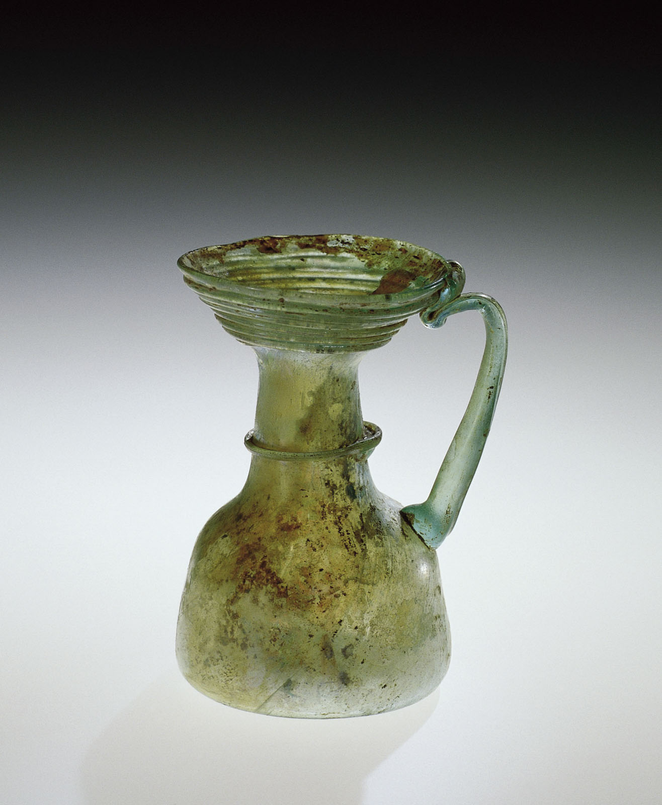 Blue-green tinted glass pitcher with a flared mouth, curving round handle, and a stout, rounded base, showing signs of aging