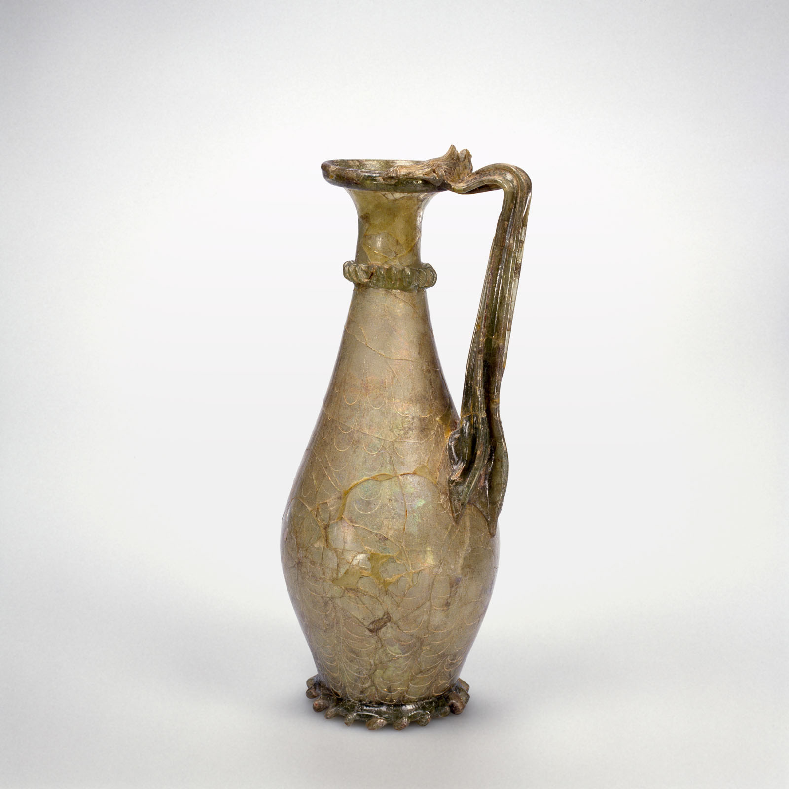 Transparent glass pitcher with a tall, slender form and a flattened handle, featuring a flared rim, a  textured body, and decorative base, showing signs of aging