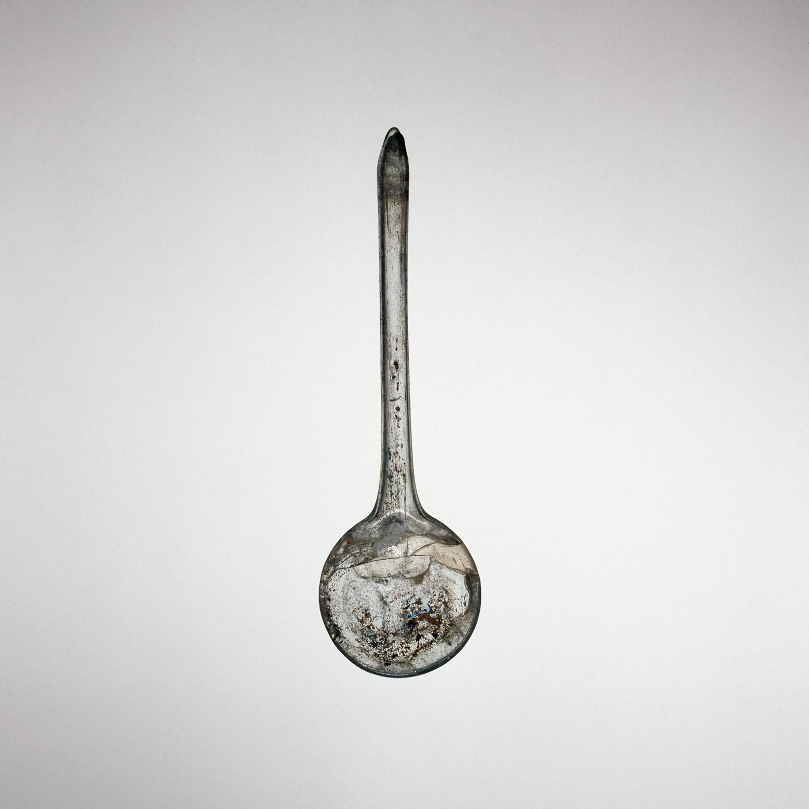 Transparent glass spoon with a long, straight handle and a deep, rounded bowl, showing evidence of aging