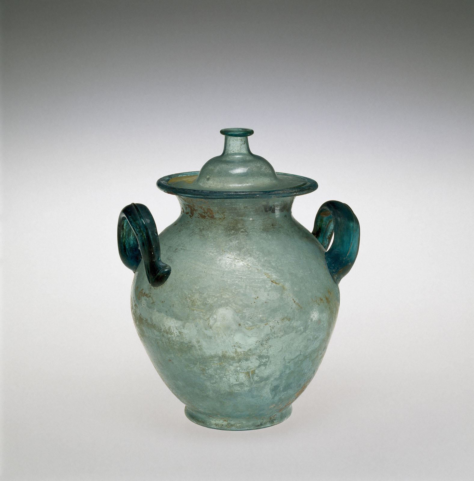 Covered glass urn in pale blue, featuring two handles and a matching lid, with a worn surface