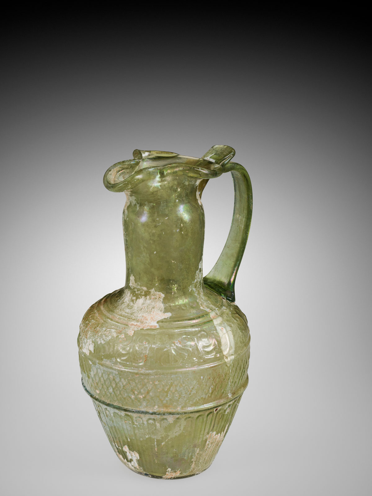 Pale yellow-green glass pitcher with a curled lip and handle, detailed with embossed designs on its body, showing extensive signs of aging