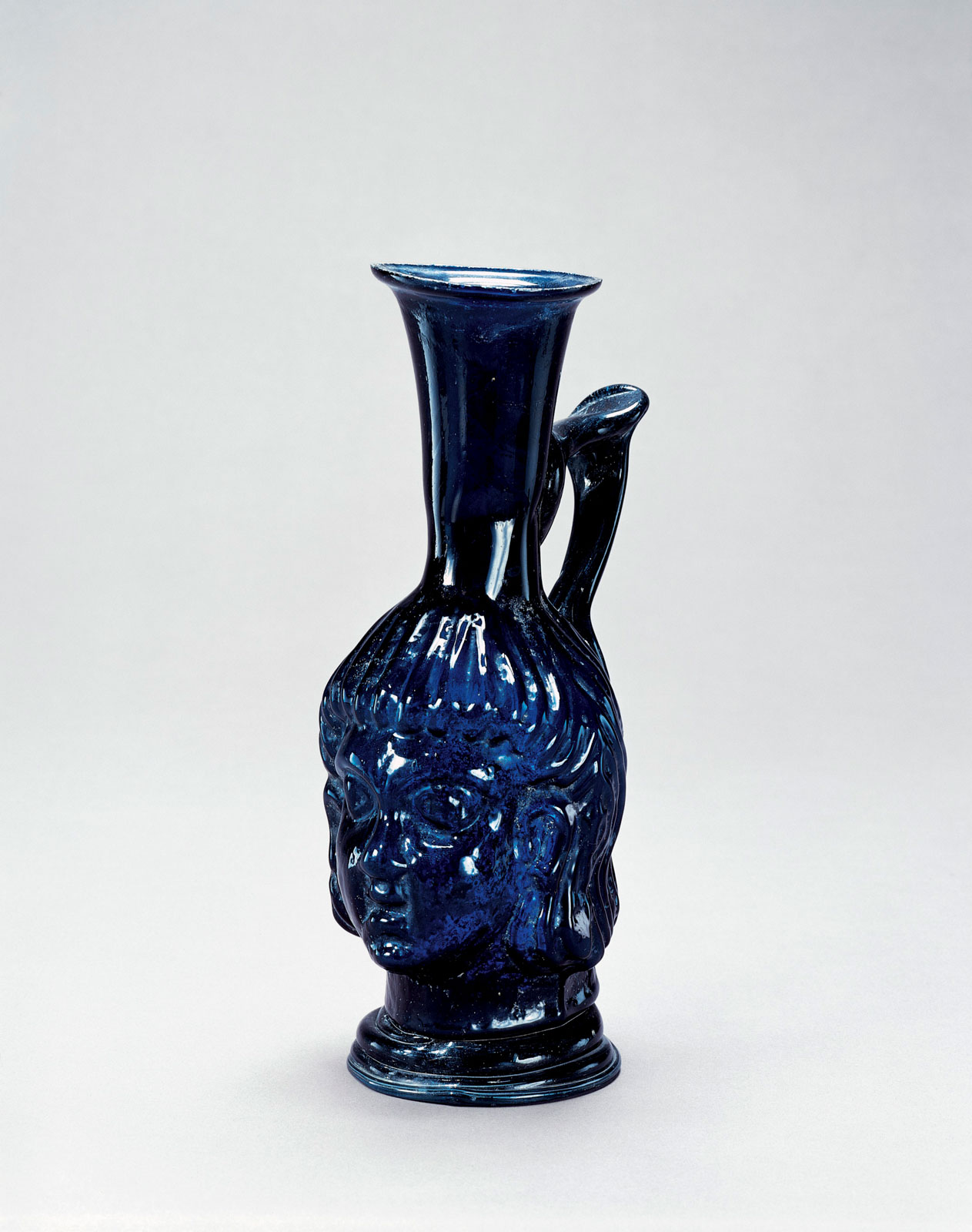 Cobalt blue glass pitcher in the form of a detailed head/face, featuring a flared neck and angled handle