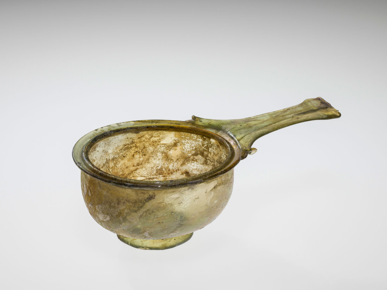 Mottled yellow-green, round, glass  bowl with a single, long, flat yellow-green handle sticking straight out, showing extensive signs of aging