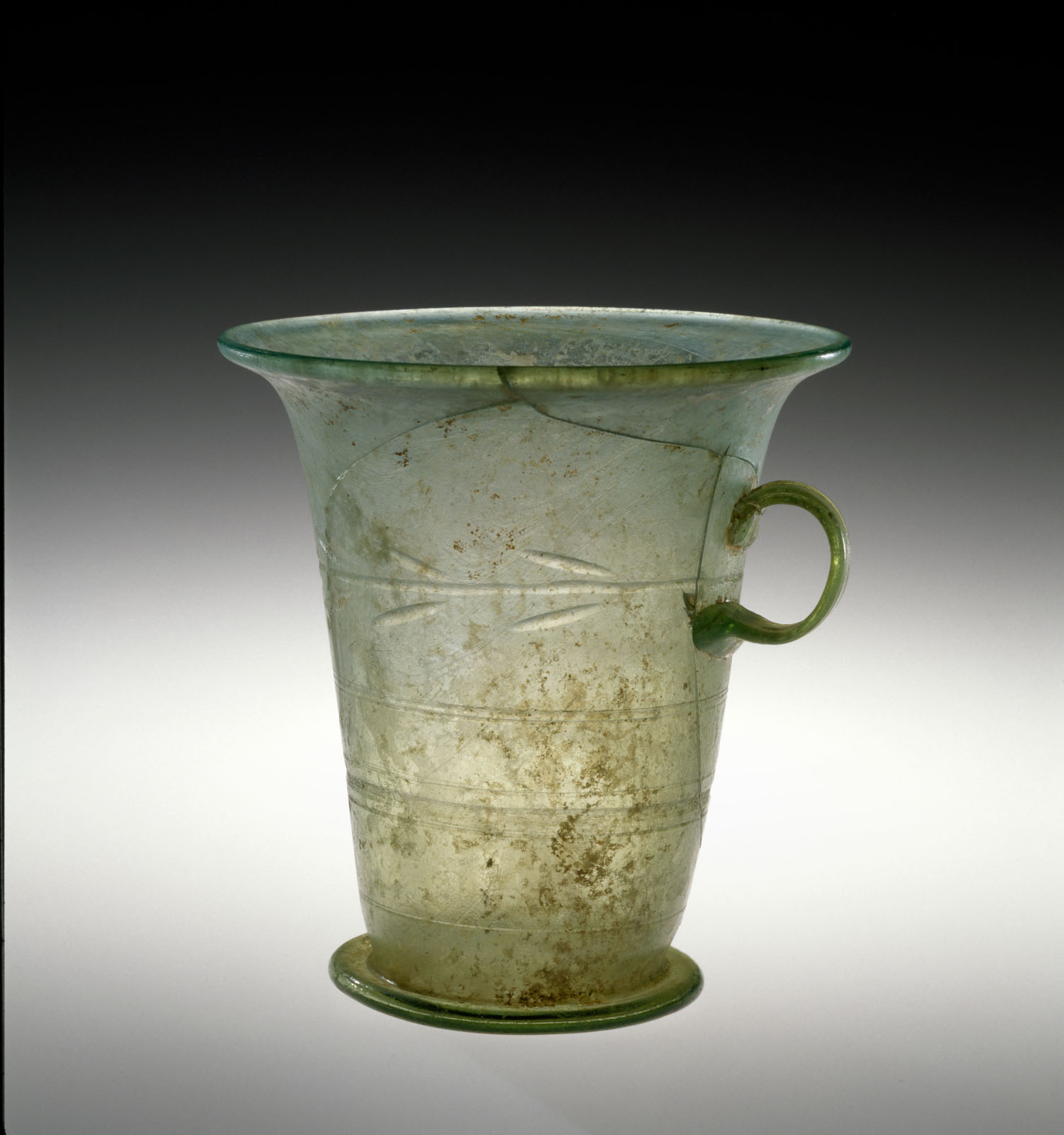 Cracked, green glass beaker with a flared rim and mall round handle, adorned with horizontal bands, showing signs of aging