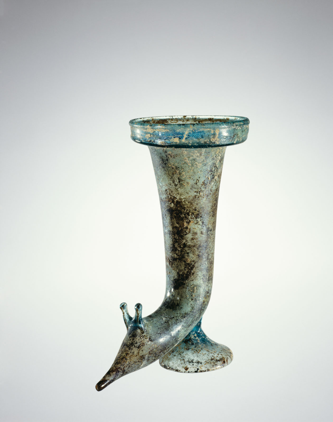 Mottled blue-green glass horn-shaped vessel with pointy-nosed animal end and flared base, showing extensive signs of aging