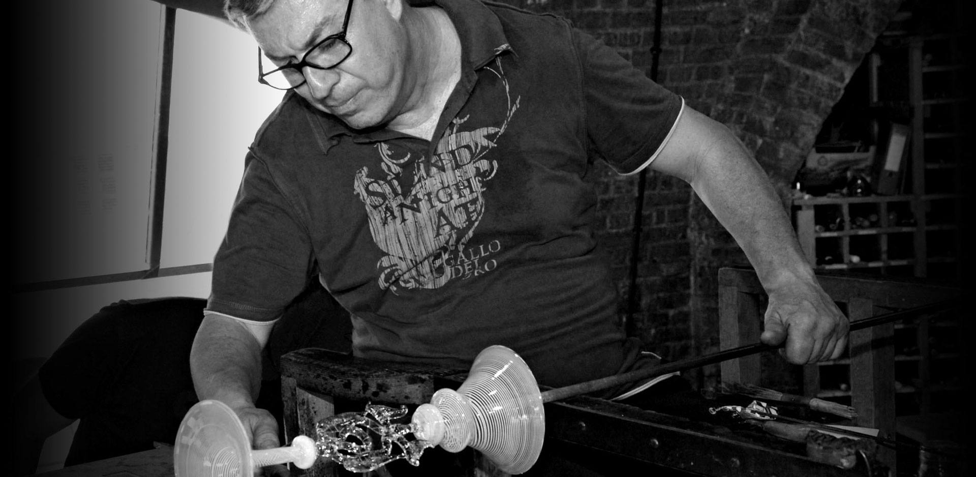 Monochrome image of a glassblower shaping an ornate glass vessel on a blowpipe